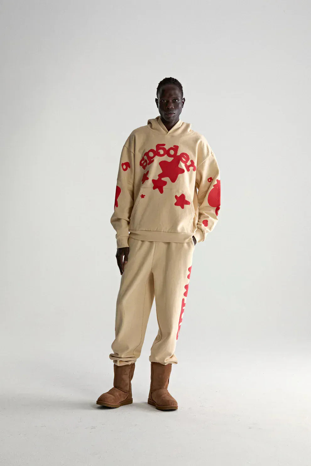 A person stands wearing the Sp5der 440GSM Heavyweight Star Hoodie in cream with red puff print—an oversized streetwear pullover—styled with joggers and brown boots against a plain light background, highlighting modern street style.