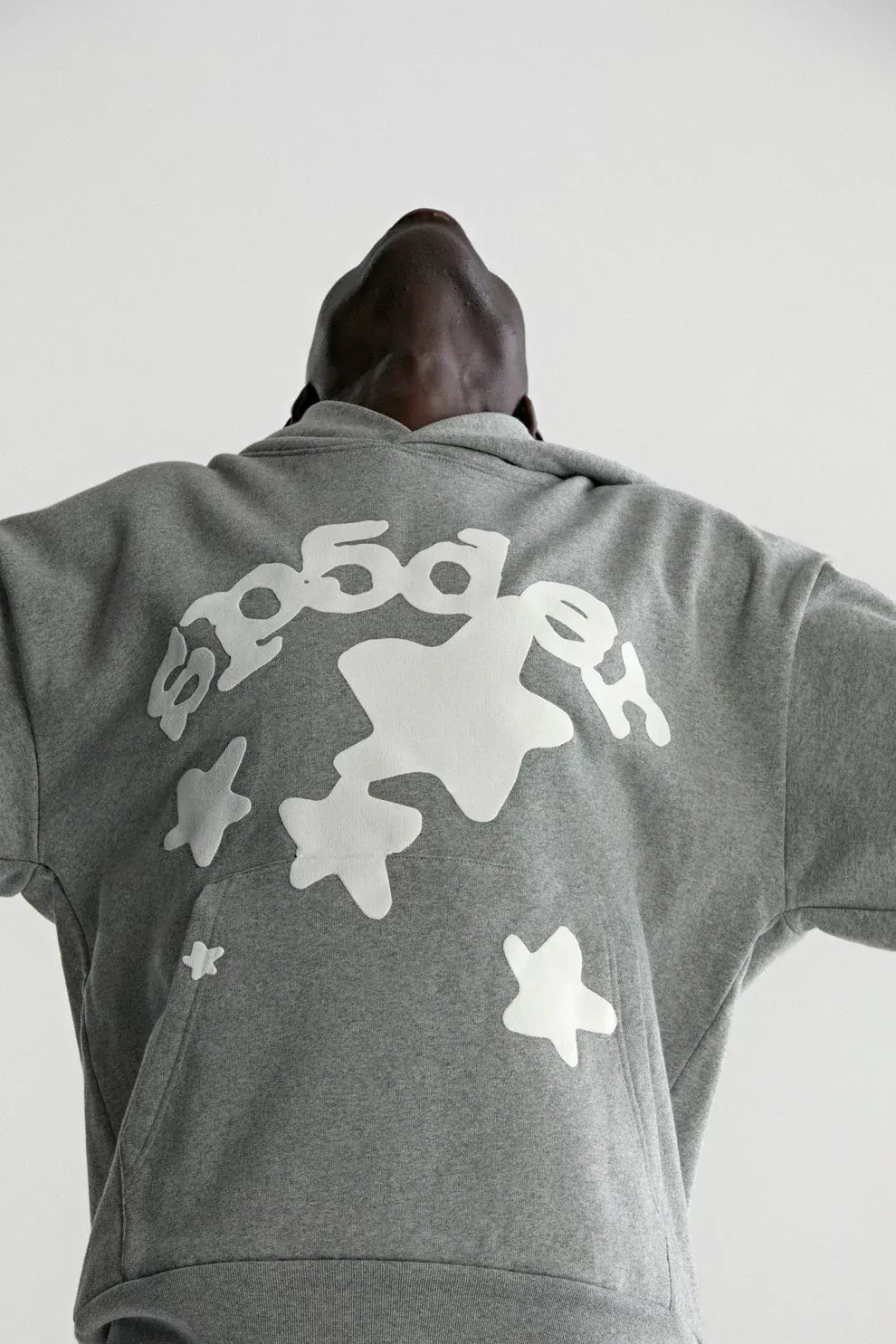 Person in a Sp5der Stars Hoodie – Grey 440GSM Heavyweight Streetwear Pullover, featuring white abstract stars and "sp5der" on the front, head tilted back, photographed from a low angle.