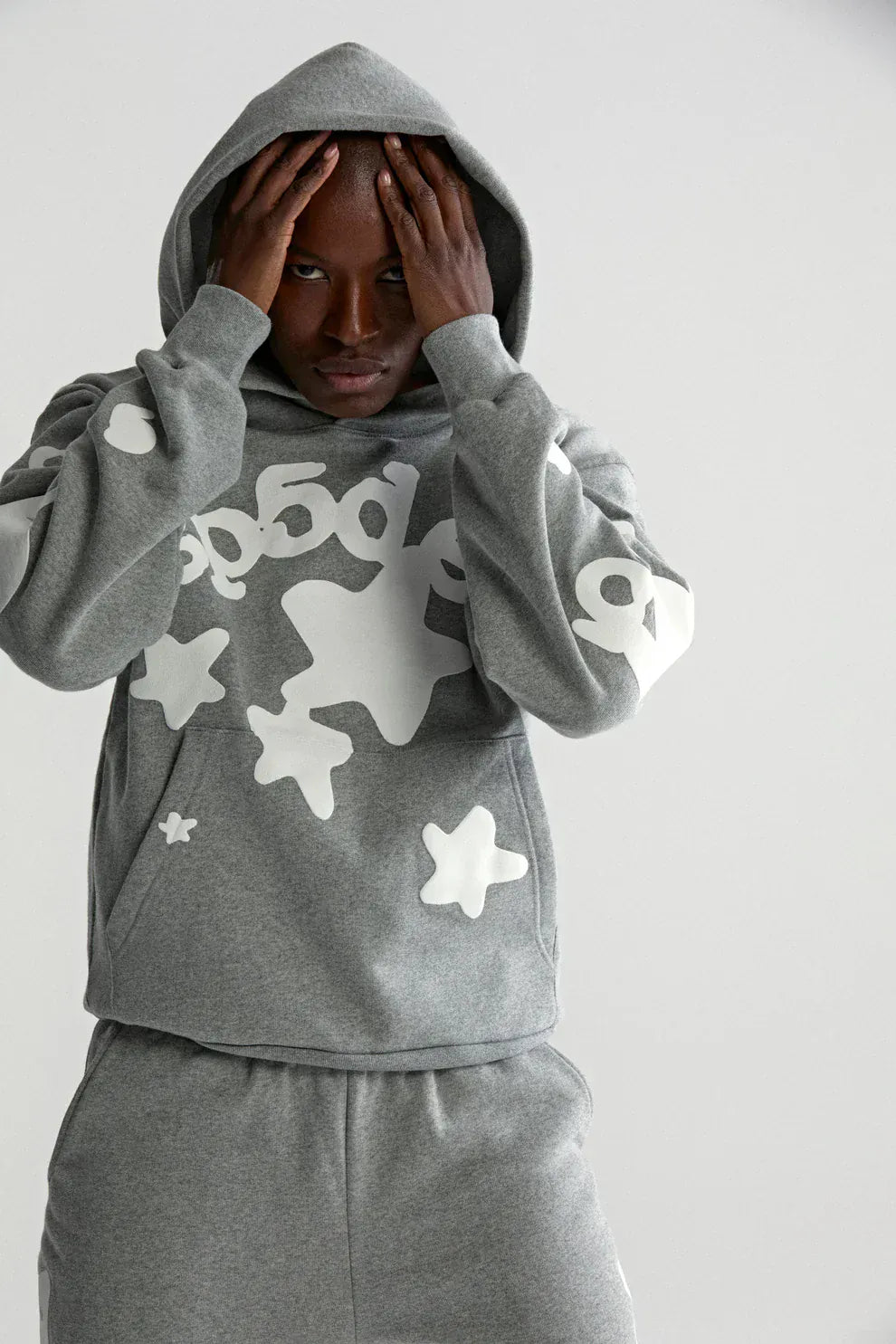 A person stands against a plain background, wearing the Sp5der Stars Hoodie – Grey 440GSM Heavyweight Streetwear Pullover with matching sweatpants featuring white abstract star and number designs, hands held to their head.
