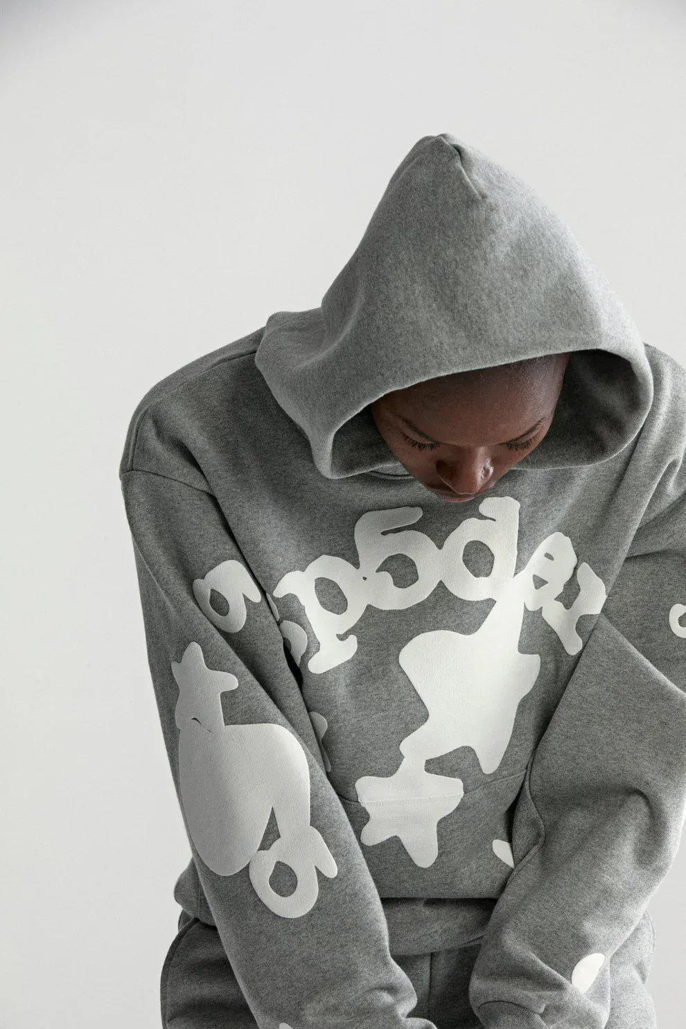 Wearing the Sp5der Stars Hoodie – Grey 440GSM Heavyweight Streetwear Pullover, a person with the hood partly over their head looks downward. The hoodie features large white graphic text and shapes on gray cotton fleece.