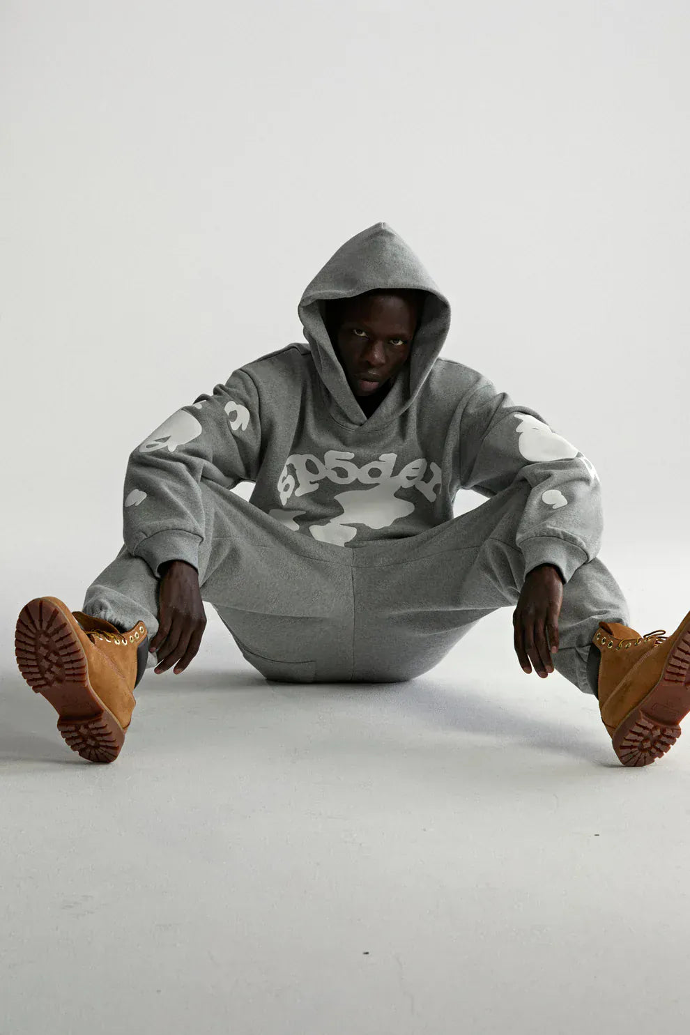 Wearing the Sp5der Stars Hoodie – Grey 440GSM Heavyweight Streetwear Pullover and sweatpants with white graphics, a person in tan boots sits on the floor with legs apart, set against a plain light background.