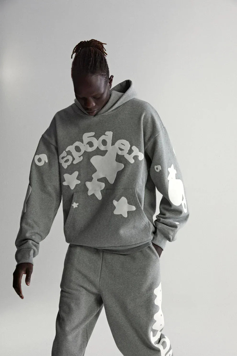 A person wears the Sp5der Stars Hoodie – Grey, a 440GSM heavyweight streetwear pullover with bold white graphics, paired with matching sweatpants, standing against a plain light background.