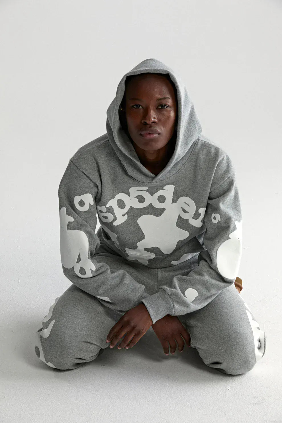A person kneels on a white surface, facing the camera, wearing the Sp5der Stars Hoodie – Grey 440GSM Heavyweight Streetwear Pullover with bold white abstract designs and matching sweatpants.