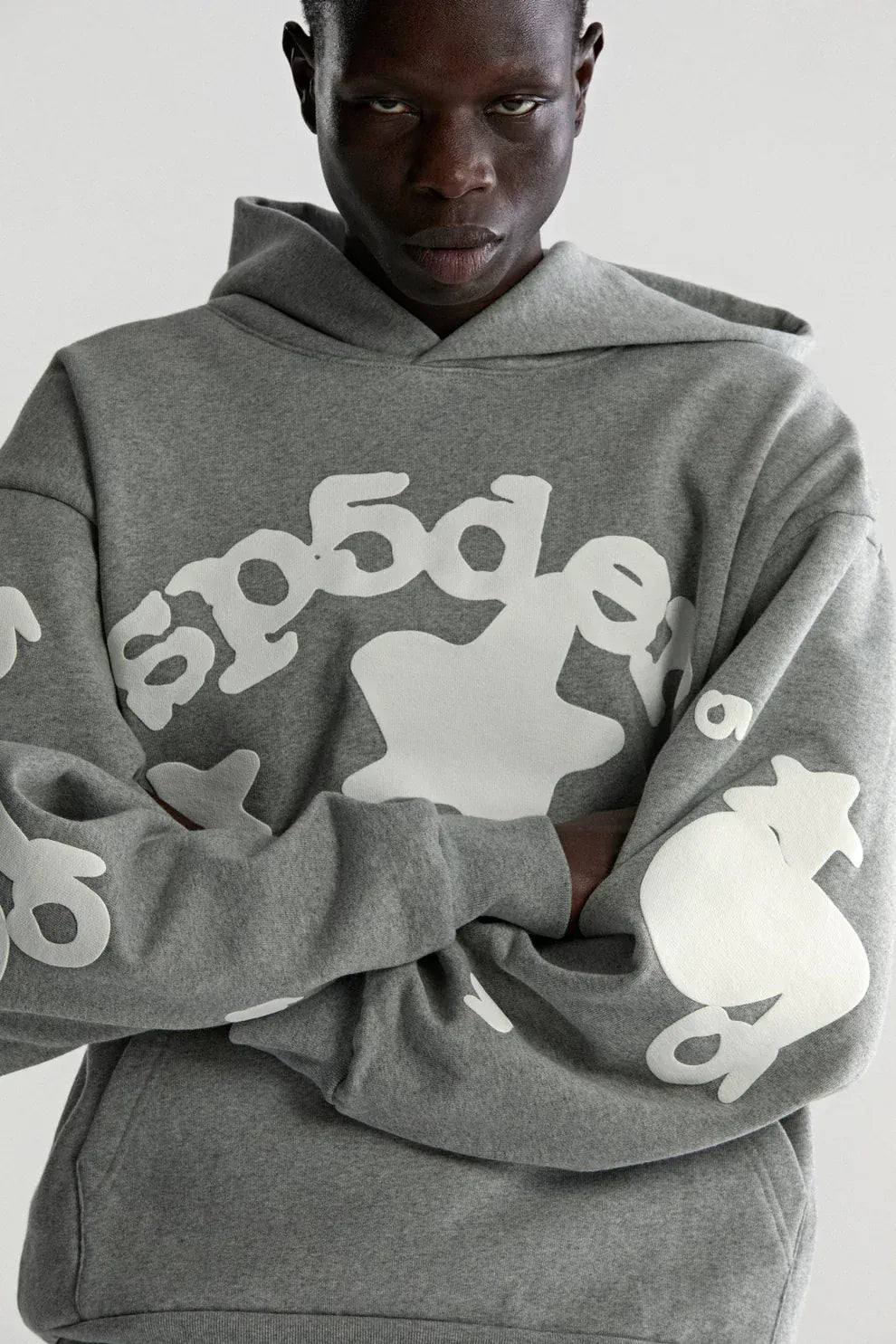 A person wearing the Sp5der Stars Hoodie – Grey 440GSM Heavyweight Streetwear Pullover with bold white graphic text and symbols on the front and sleeves stands with arms crossed against a plain background.