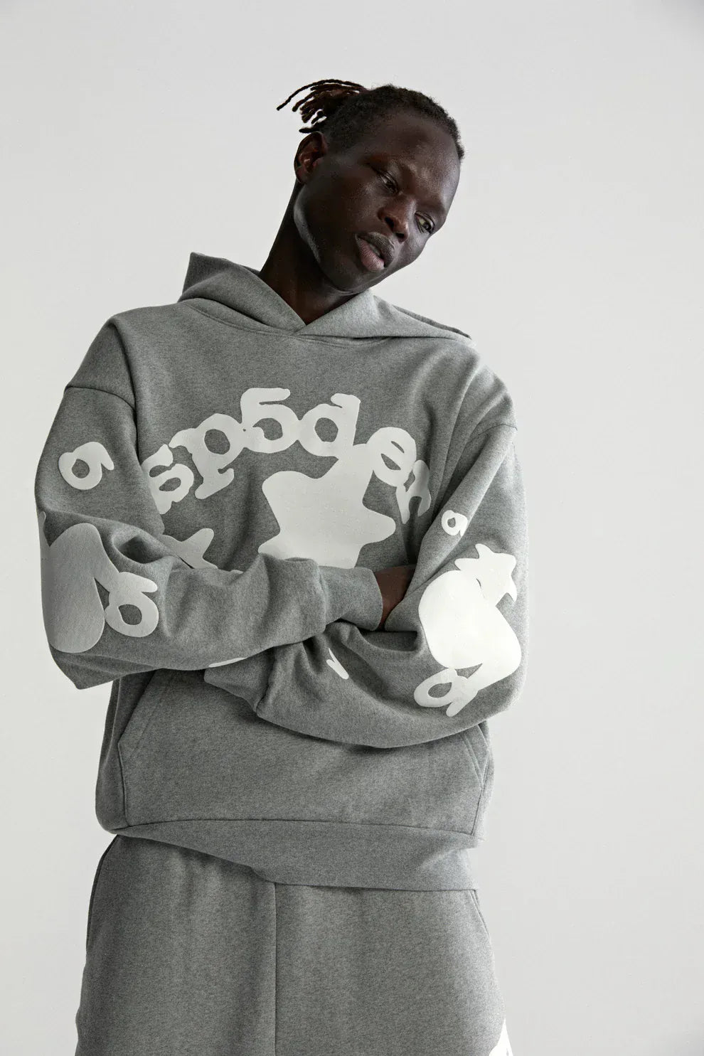 A person wears the Sp5der Stars Hoodie – Grey 440GSM Heavyweight Streetwear Pullover with matching pants, featuring abstract white text and shapes, standing against a plain light background.