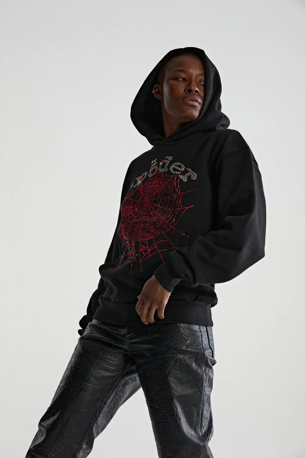 Wearing the Sp5der Rhinestone Web Hoodie – Black 440GSM Heavyweight Oversized Streetwear Pullover with black textured pants, a person stands against a plain background, showcasing luxury streetwear style with its red spiderweb design.
