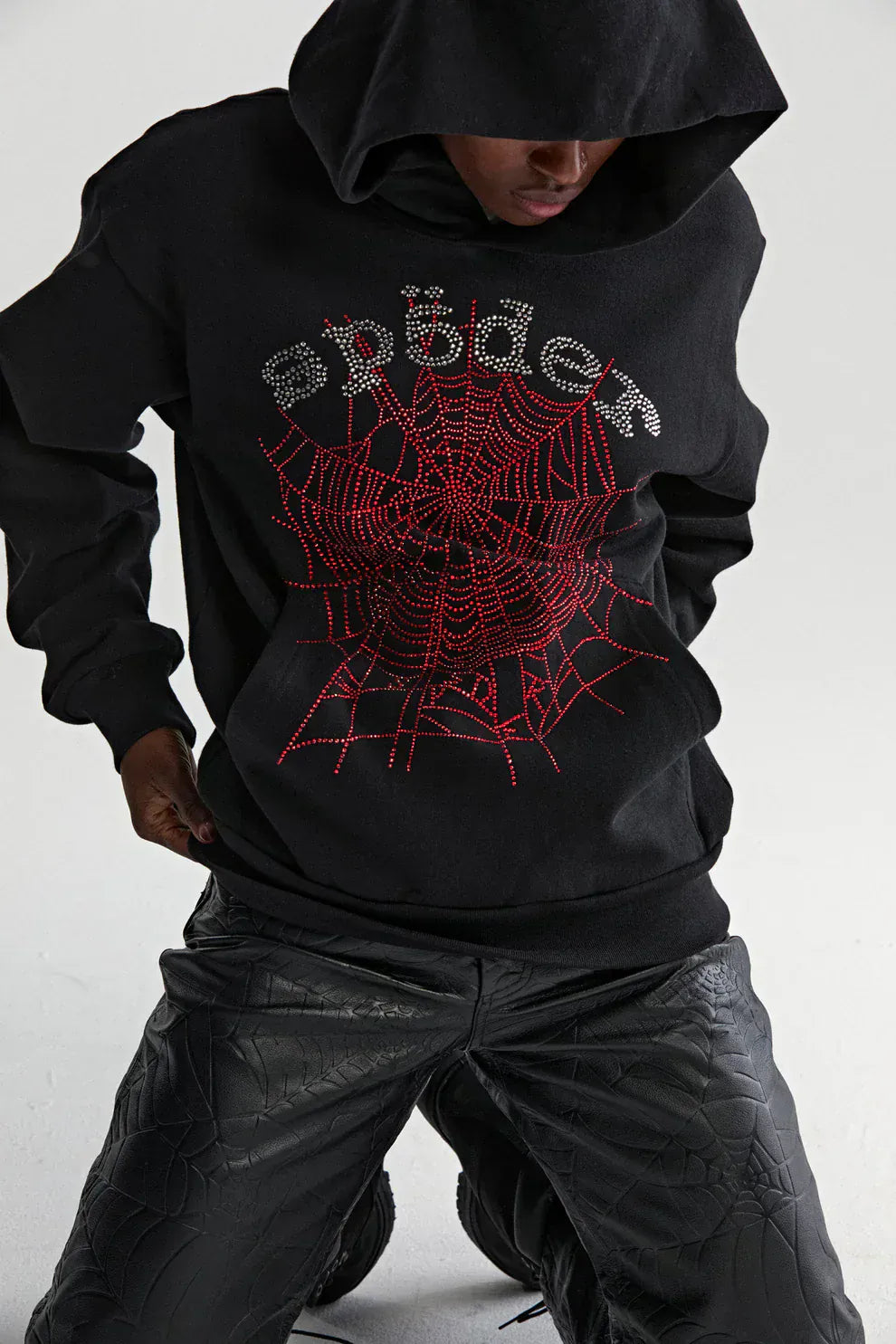 A person kneels on a plain background wearing the Sp5der Rhinestone Web Hoodie in black, crafted from 440GSM heavyweight cotton and featuring rhinestone spiderweb details, paired with black textured pants—ideal for luxury streetwear fans.