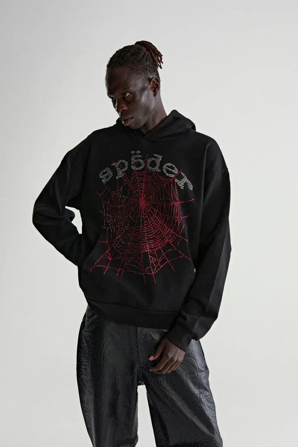 Wearing the Sp5der Rhinestone Web Hoodie – Black 440GSM Heavyweight Oversized Streetwear Pullover, paired with black textured pants, showcases luxury street style against a plain background.