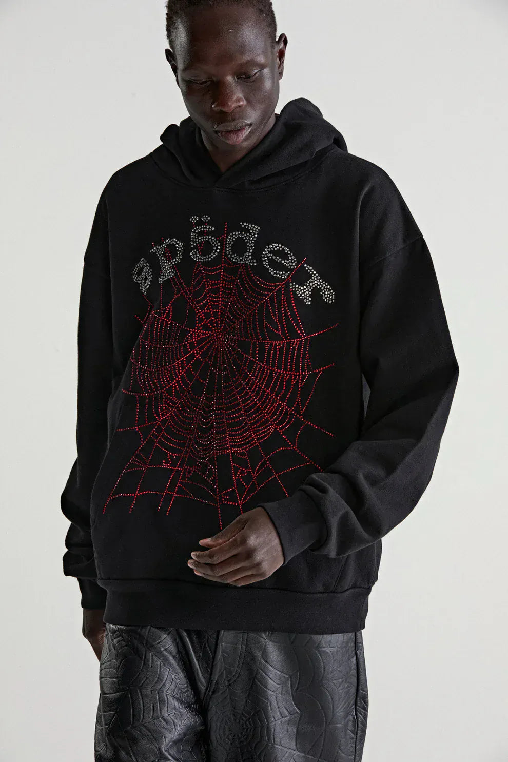 Someone wears a Sp5der Rhinestone Web Hoodie – Black 440GSM Heavyweight Oversized Streetwear Pullover, detailed with rhinestone spider and red web, paired with black patterned pants for the ideal luxury streetwear look.