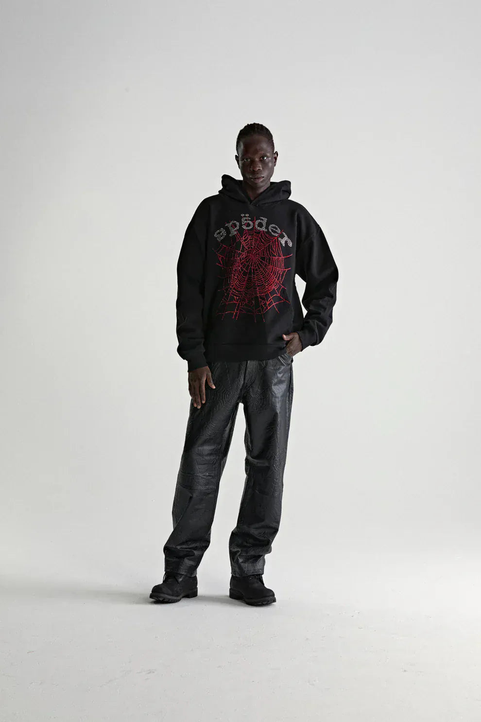 A person stands against a plain background wearing a Sp5der Rhinestone Web Hoodie – Black 440GSM Heavyweight Oversized Streetwear Pullover, paired with black textured pants and shoes—a sleek luxury streetwear look.