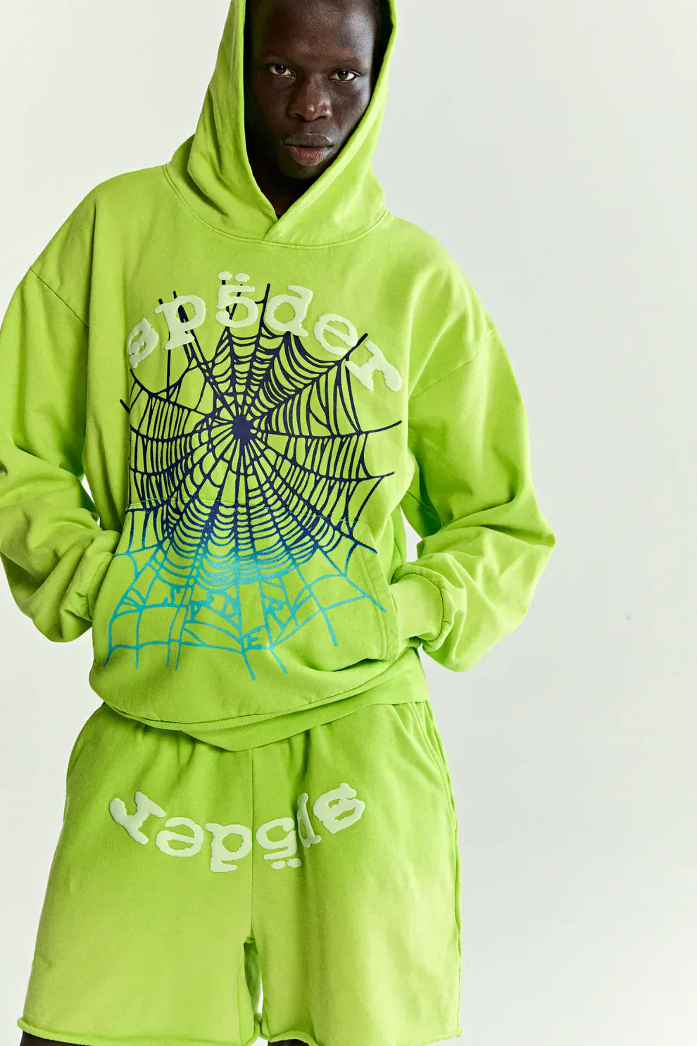 Wearing the Sp5der Web Graphic Hoodie – Neon Green 440GSM Heavyweight Streetwear, paired with matching shorts featuring bold spider text and a large black web graphic, they capture standout style against a plain background.