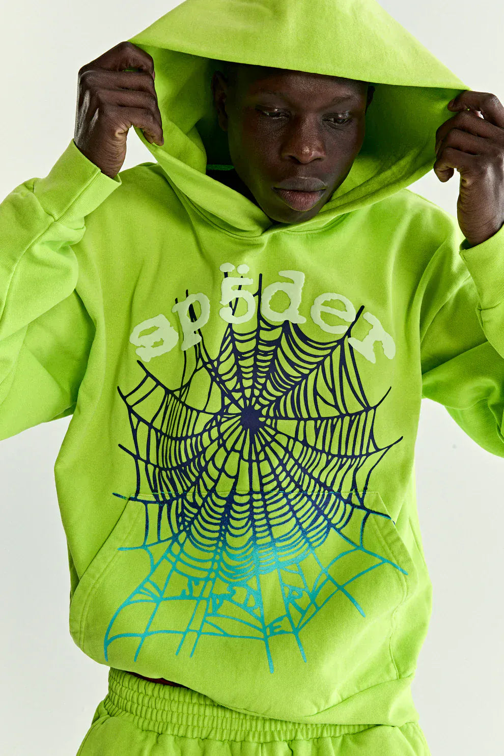 A person adjusts the hood of a Sp5der Web Graphic Hoodie – Neon Green 440GSM Heavyweight Streetwear, which features a bold black spiderweb and “sp5der” printed on the front.