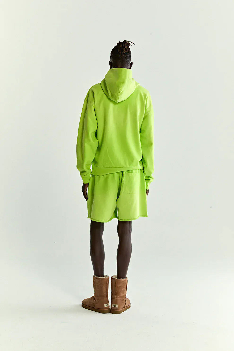 A person stands facing away in the Sp5der Web Graphic Hoodie – Neon Green 440GSM Heavyweight Streetwear with matching shorts and brown boots, set against a plain light background.