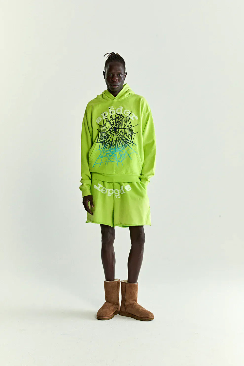 A person stands against a plain background wearing a Sp5der Web Graphic Hoodie – Neon Green 440GSM Heavyweight Streetwear, matching shorts, and brown boots.