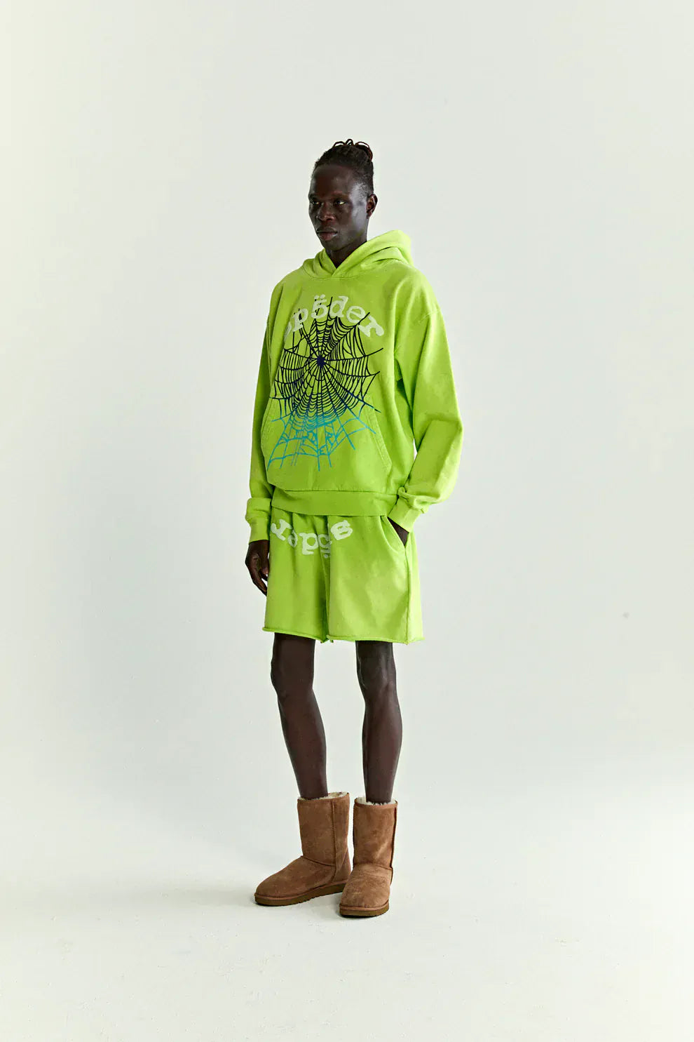 Wearing the Sp5der Web Graphic Hoodie – Neon Green 440GSM Heavyweight Streetwear with matching shorts and brown shearling boots, a person stands against a plain light background for a striking streetwear look.