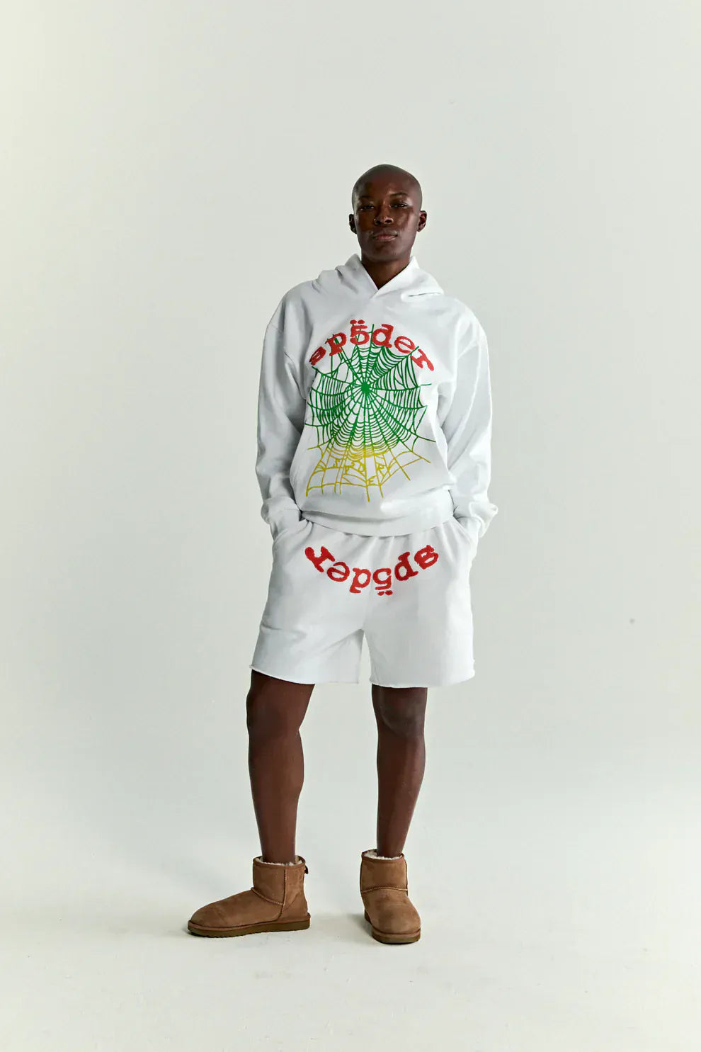 A person wears the Sp5der “Toxic Web” Hoodie in white & neon lime, a 440GSM heavyweight oversized streetwear pullover with a colorful web graphic, styled with shorts and brown ankle boots against a plain background.