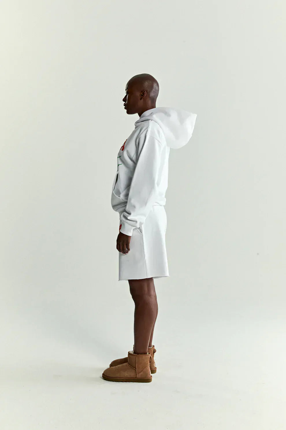 A person stands in profile against a plain background, wearing a white & neon lime Sp5der “Toxic Web” Hoodie 440GSM Heavyweight Oversized Streetwear Pullover, white shorts, and tan ankle boots for a modern Y2K streetwear vibe.