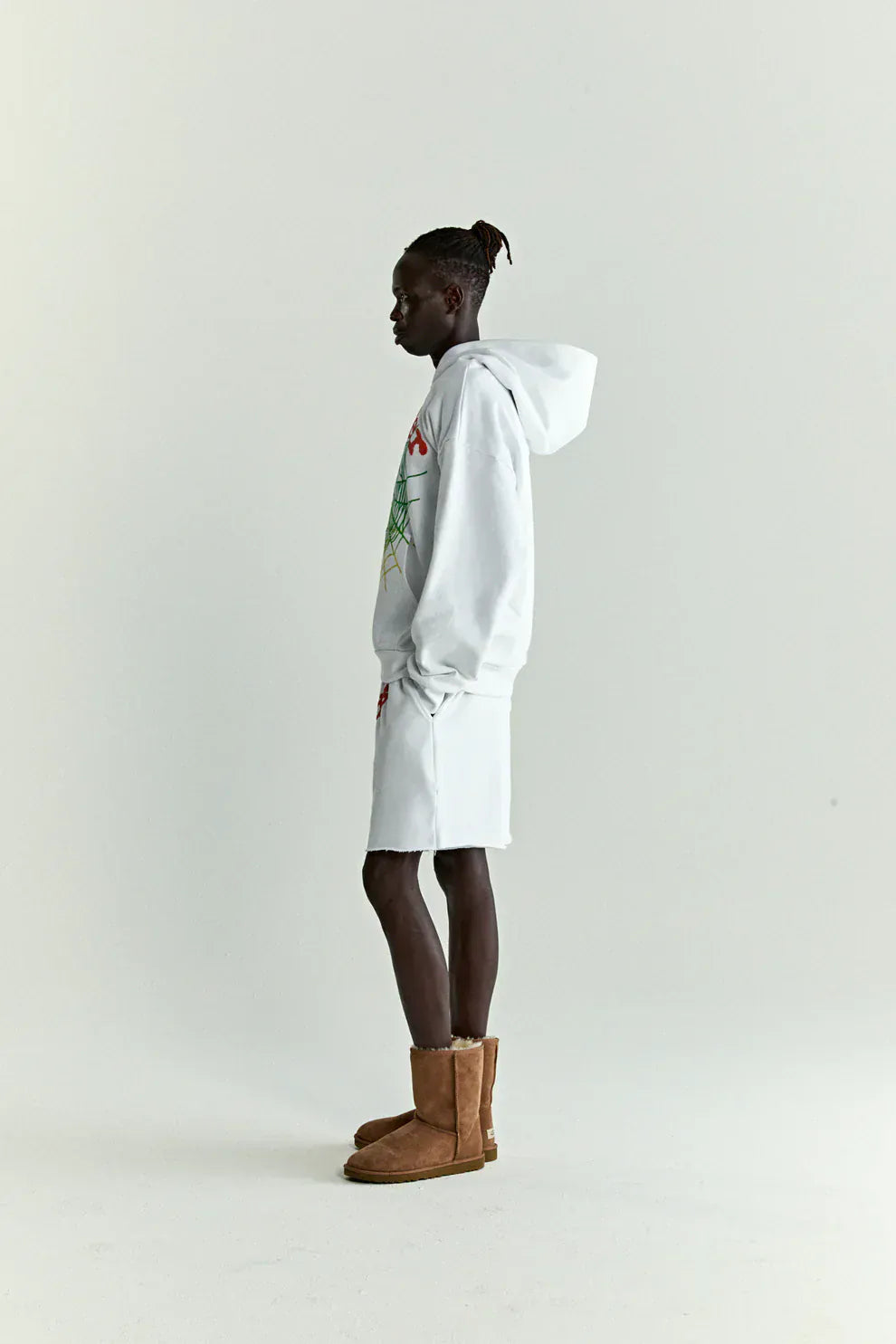 A person stands in profile against a plain background, wearing a Sp5der “Toxic Web” Hoodie in White & Neon Lime—an oversized 440GSM heavyweight streetwear pullover—paired with white shorts and brown boots for a Y2K-inspired look.