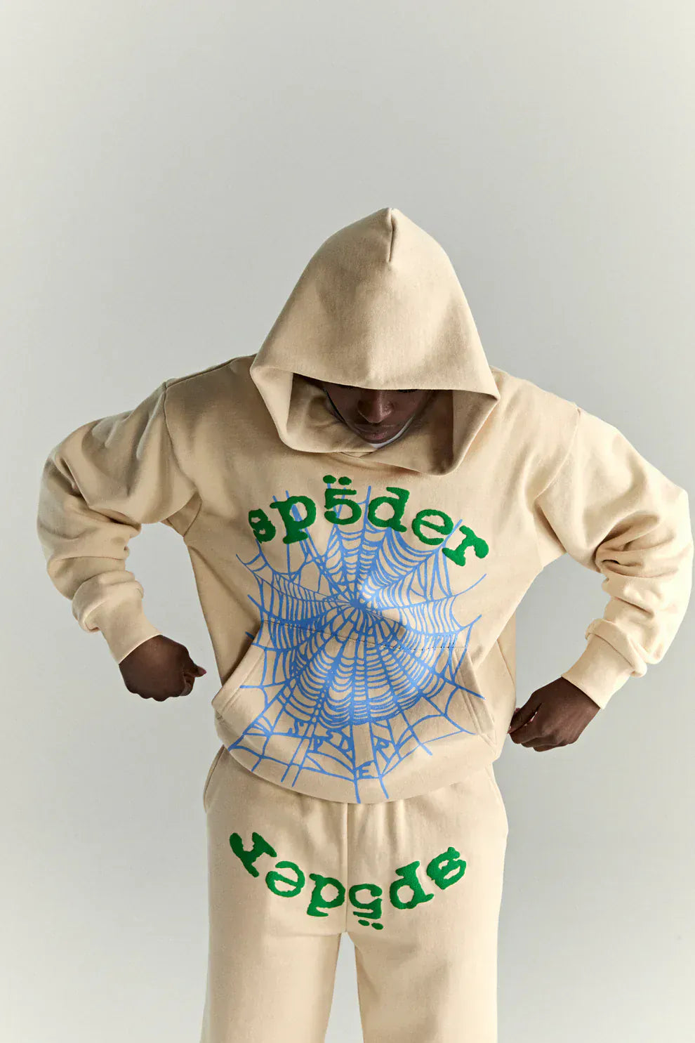 Person wearing a Sp5der “Sky Web” Hoodie in cream with blue and green details, 440GSM oversized streetwear pullover, both hands on hips and head down, showcasing bold Y2K vibes and graphic spiderweb design.