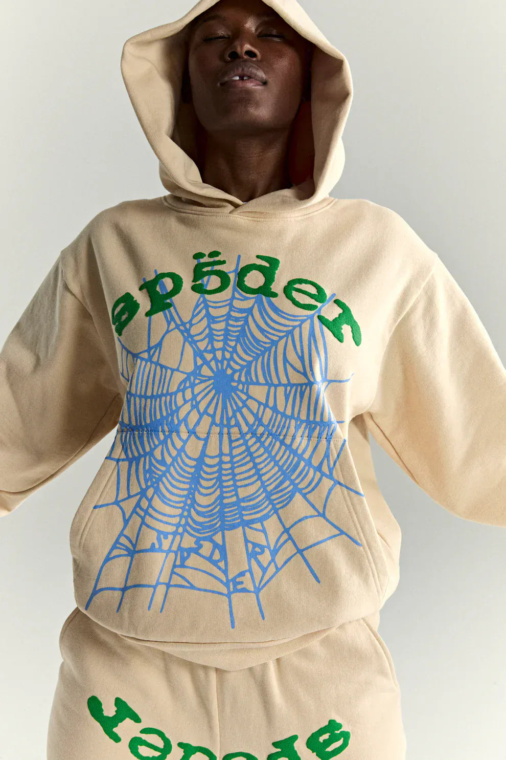 Person in a Sp5der “Sky Web” Hoodie—Cream/Blue/Green 440GSM Oversized Streetwear Pullover—featuring bold web graphics and green text, makes the perfect Y2K statement with arms slightly raised.