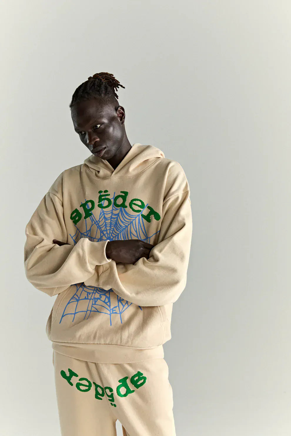 Dressed in a Sp5der “Sky Web” Hoodie – Cream / Blue / Green 440GSM Oversized Streetwear Pullover, featuring blue spiderweb graphics and Y2K style, a person stands against a plain light background.