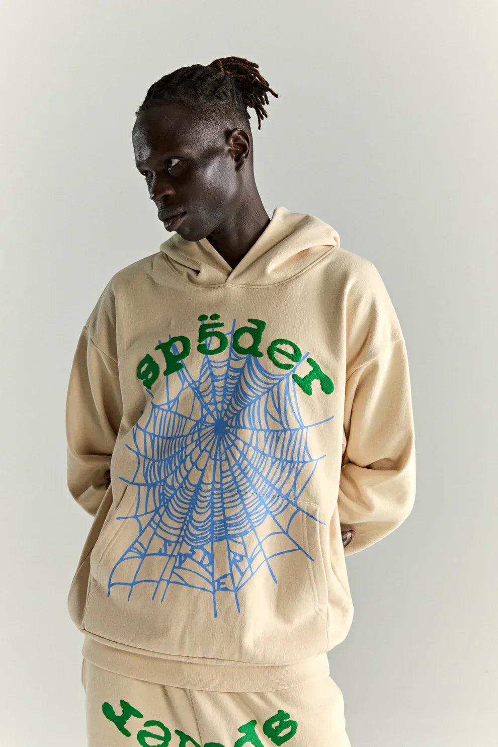 Wearing the Sp5der “Sky Web” Hoodie – Cream / Blue / Green 440GSM Oversized Streetwear Pullover, featuring a large blue spider web and green "spøder" text, a person stands against a plain light background.