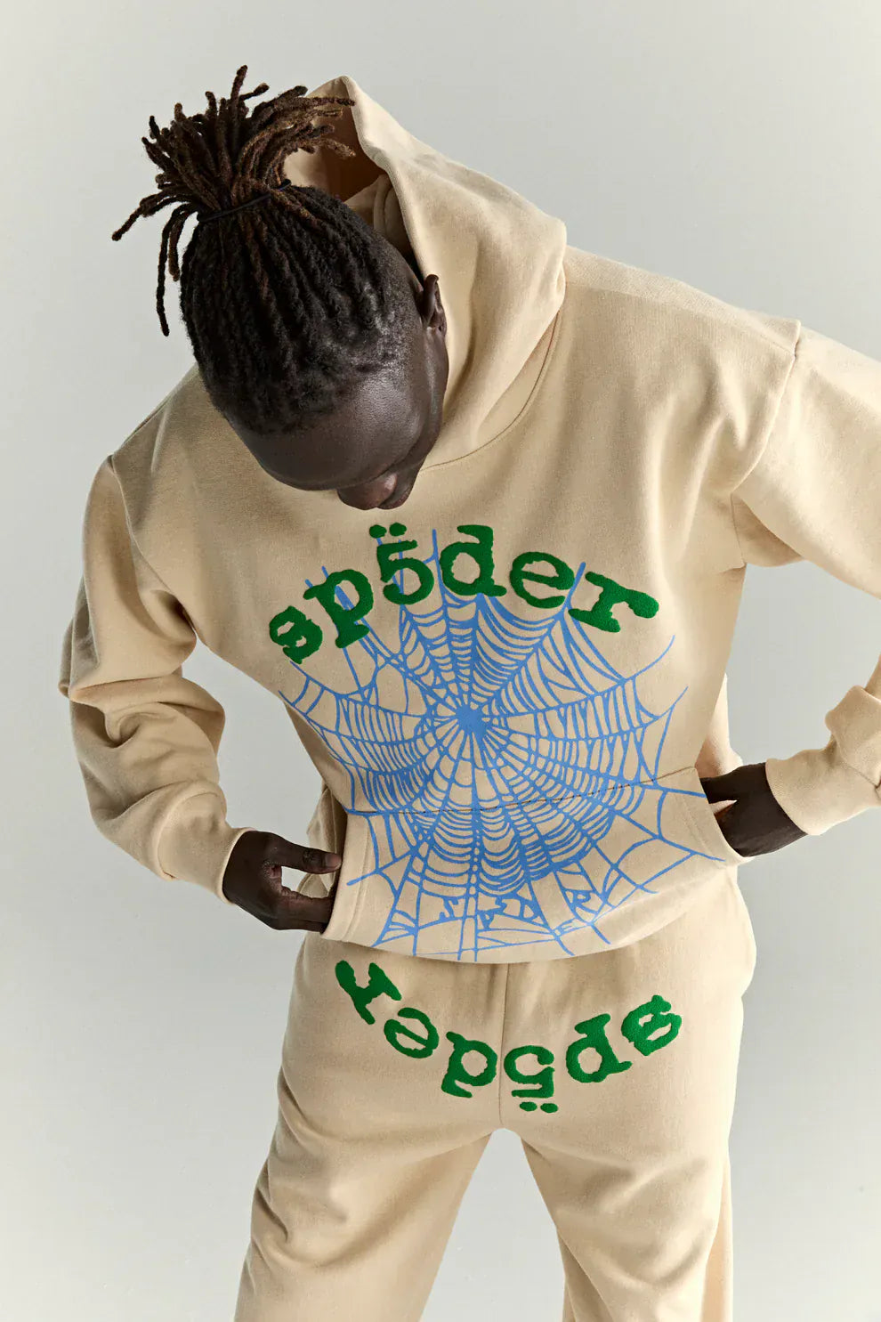 Someone wears the Sp5der “Sky Web” Hoodie in cream with blue and green details, a 440GSM heavyweight oversized streetwear pullover, paired with matching pants for a bold Y2K vibe.