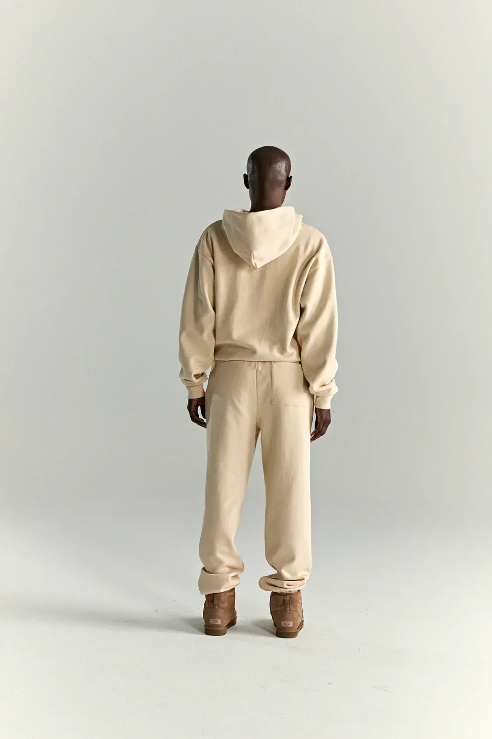 A person stands facing away, wearing the Sp5der “Sky Web” Hoodie in cream, a 440GSM oversized streetwear pullover with matching sweatpants and brown shoes, channeling Y2K vibes against a plain light background.