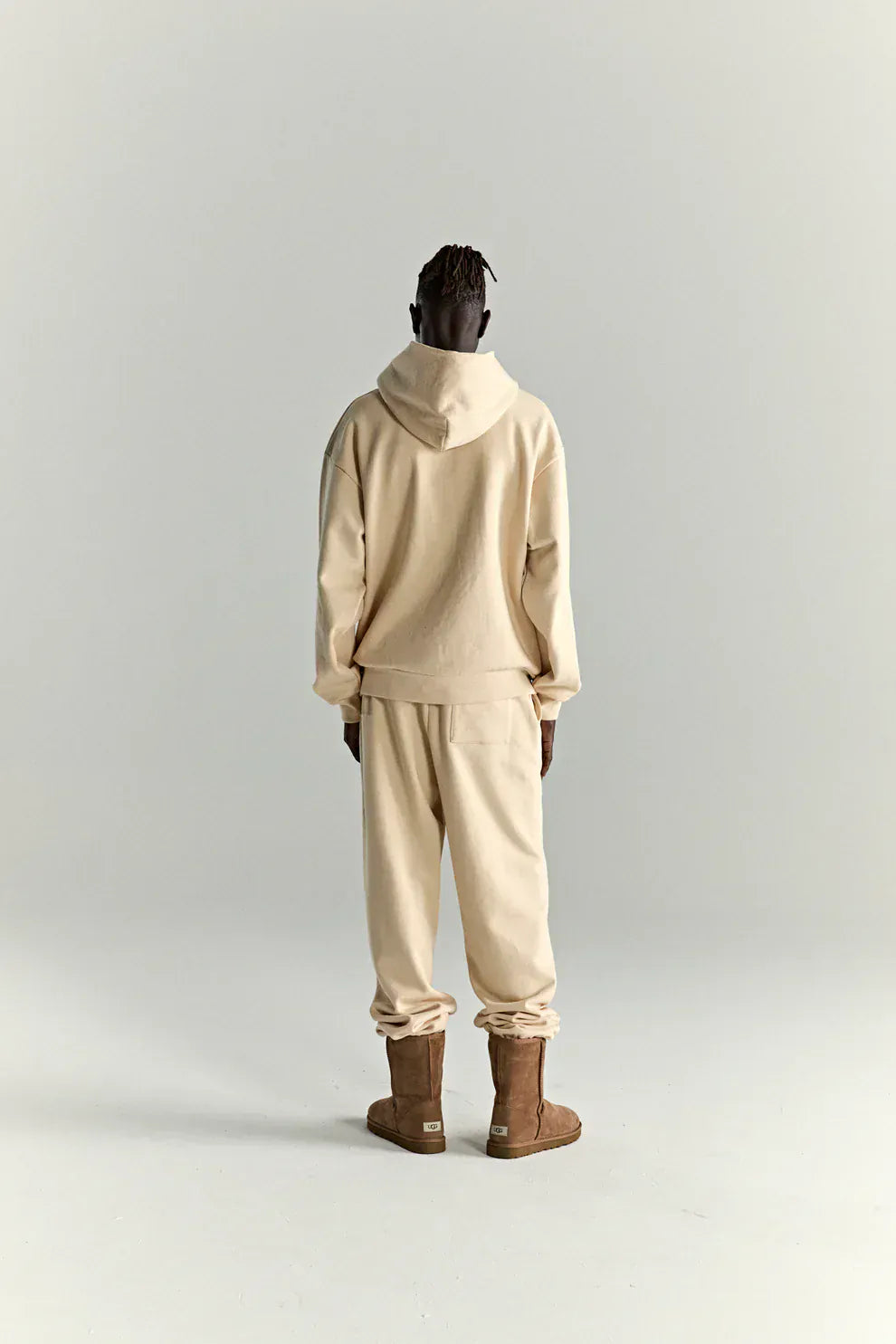 A person stands with their back to the camera, wearing a Sp5der “Sky Web” Hoodie – Cream / Blue / Green 440GSM Oversized Streetwear Pullover, matching sweatpants, and brown boots against a plain, light background.