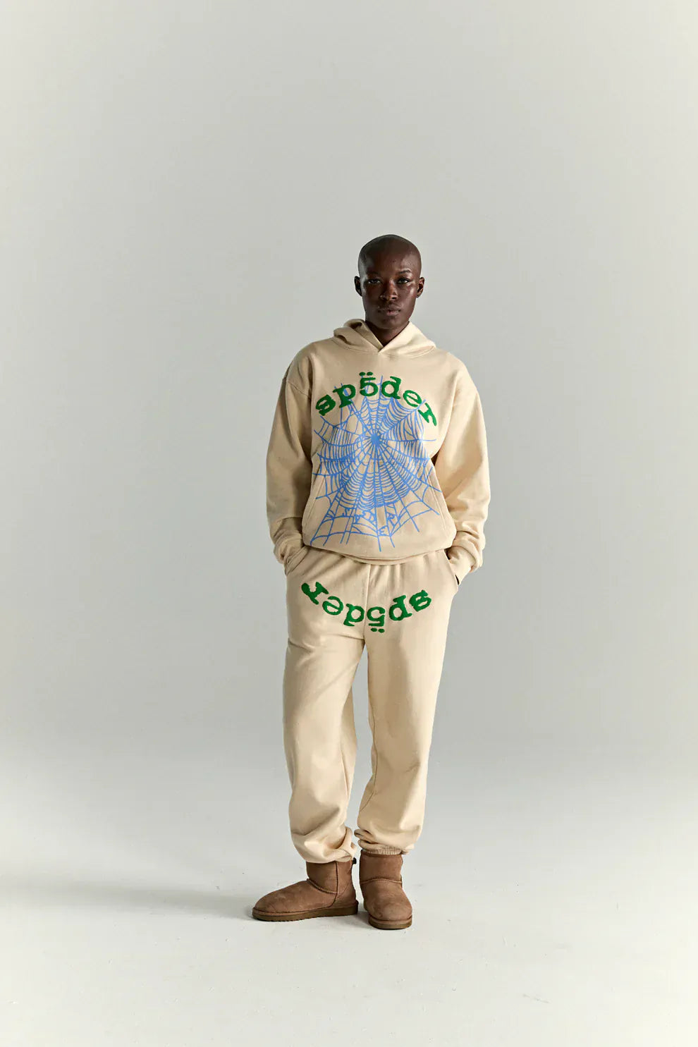 A person wears a Sp5der “Sky Web” Hoodie in cream, blue, and green (440GSM oversized streetwear pullover) with matching sweatpants and brown boots, standing against a plain light background.