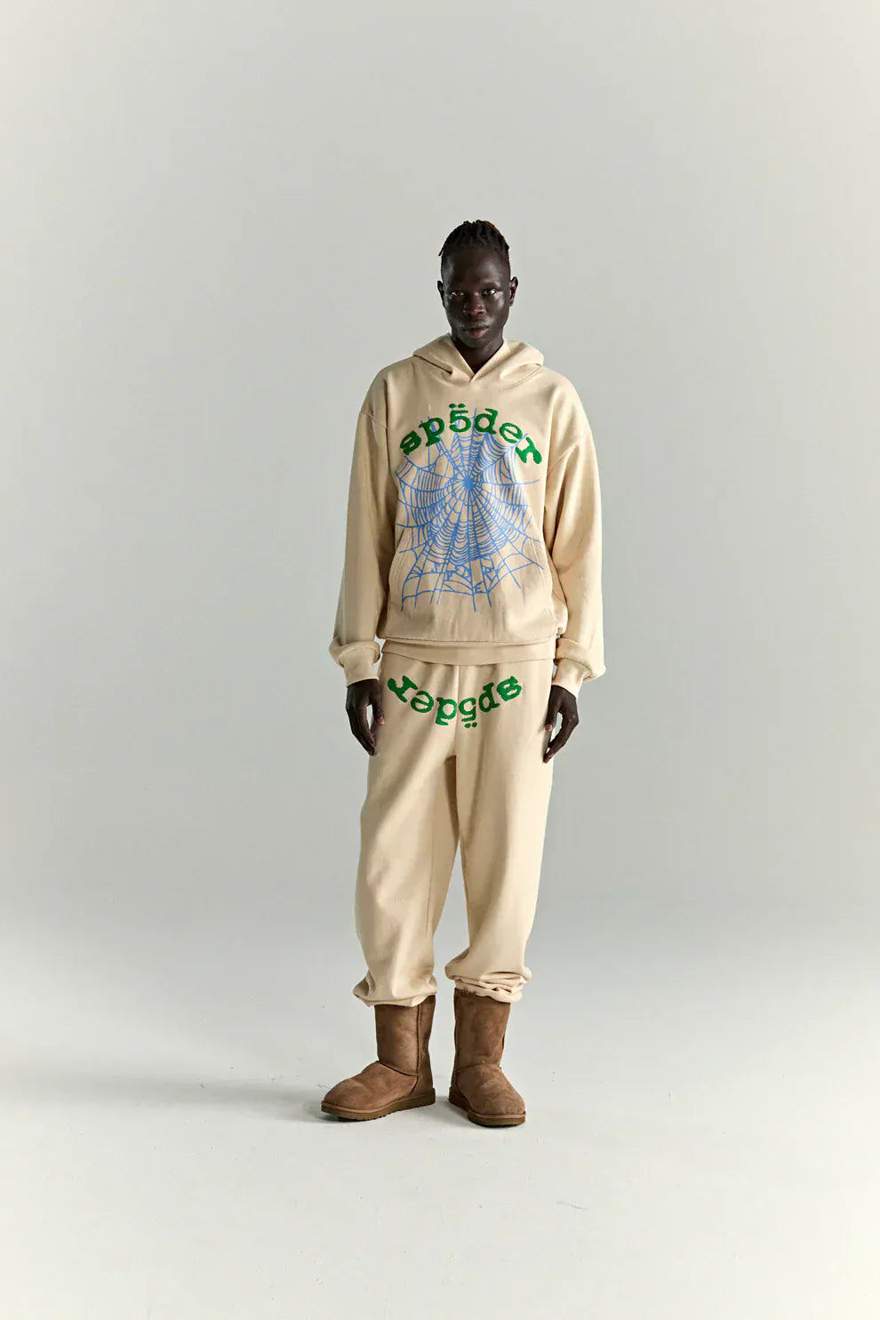 A person wears the Sp5der “Sky Web” Hoodie – Cream / Blue / Green 440GSM Oversized Streetwear Pullover with matching sweatpants and brown boots, showing off Y2K streetwear vibes against a plain light background.