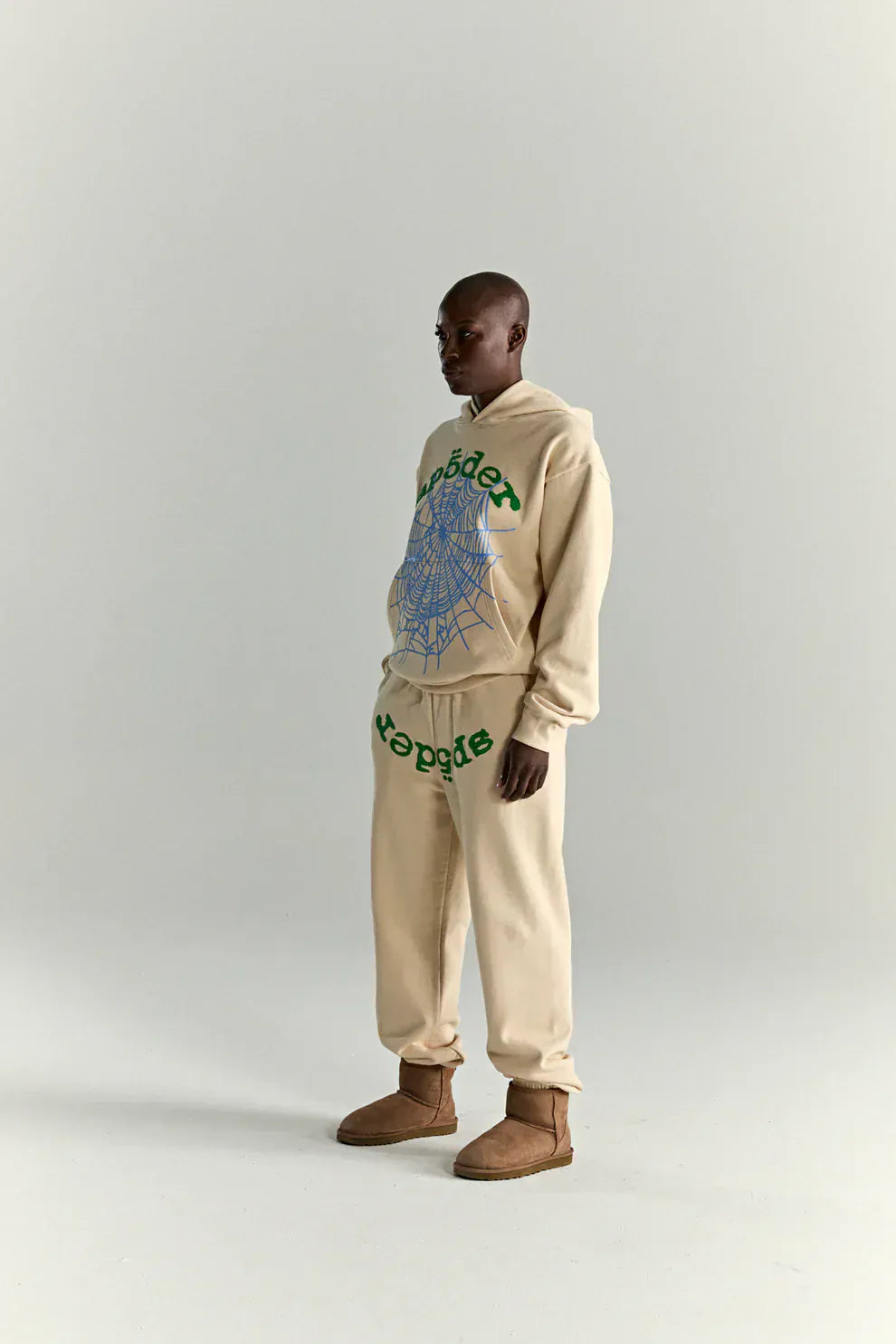 A person stands in profile wearing the Sp5der “Sky Web” Hoodie in cream with blue and green graphics (440GSM oversized streetwear pullover), paired with beige boots, giving off a casual Y2K vibe against a plain light background.