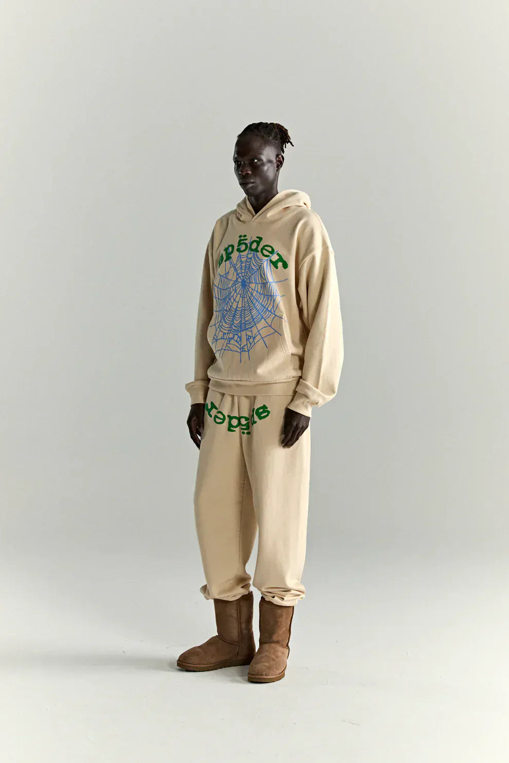 A person wears the Sp5der “Sky Web” Hoodie in cream with blue and green graphics, a heavyweight 440GSM oversized streetwear pullover, paired with matching sweatpants and brown boots, channeling Y2K vibes against a plain background.