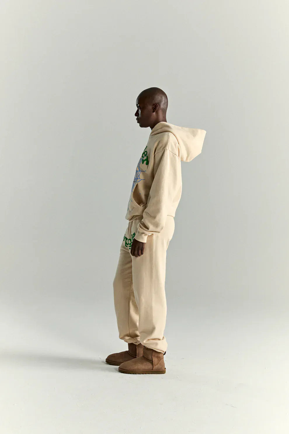A person stands in profile against a plain light background, wearing a cream Sp5der “Sky Web” Hoodie (440GSM oversized streetwear pullover), matching sweatpants, and brown slippers.