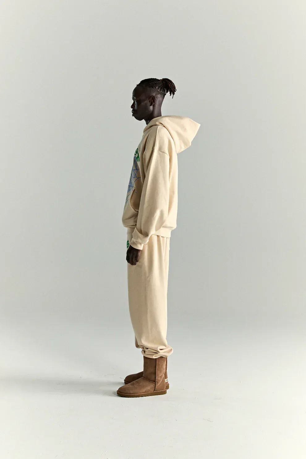 A person stands in profile wearing a Sp5der “Sky Web” Hoodie – Cream/Blue/Green 440GSM Oversized Streetwear Pullover, paired with matching sweatpants and tan boots, exuding Y2K streetwear style against a plain light background.