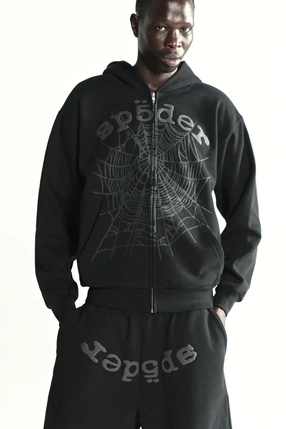 A person in the Sp5der Tonal Web Zip Hoodie – Black 440GSM Heavyweight Streetwear and matching pants, featuring "sp5der" lettering and a bold spiderweb graphic, styled as streetwear essentials.