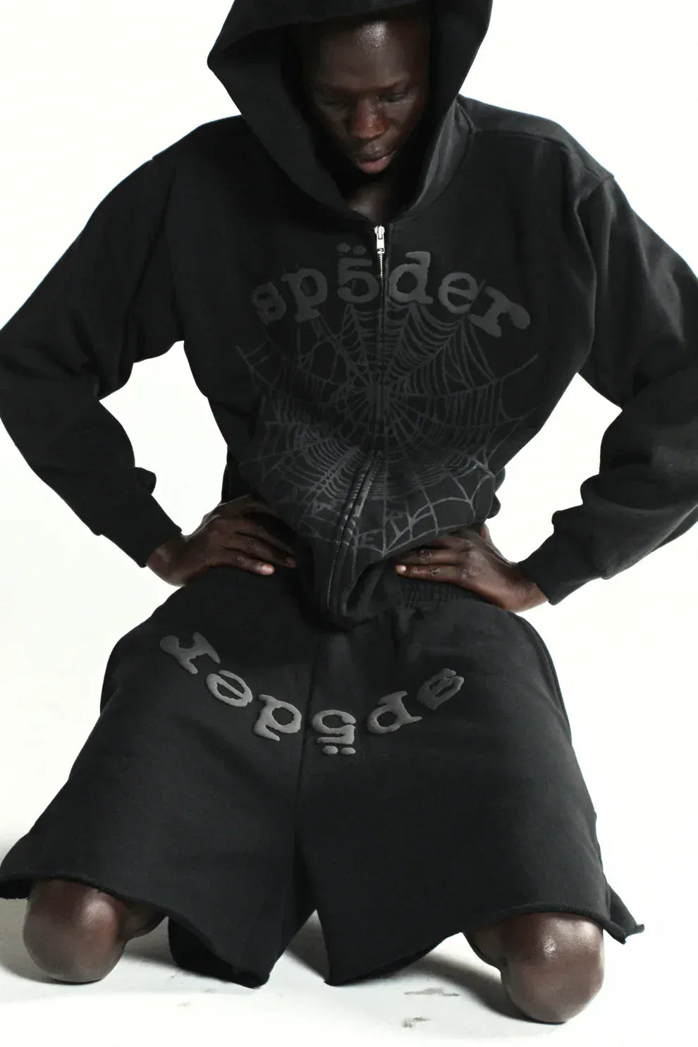 Person wearing a Sp5der Tonal Web Zip Hoodie – Black 440GSM Heavyweight Streetwear and spider web shorts, kneeling on the ground against a plain white background. This hoodie features bold web graphics for an urban edge.