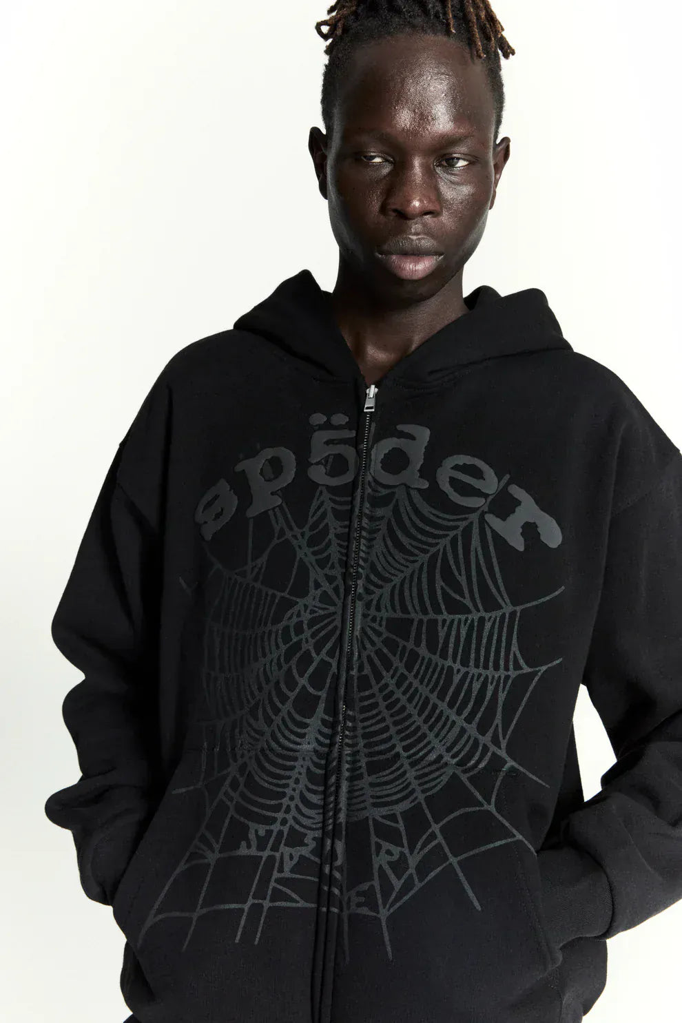 Person wearing a Sp5der Tonal Web Zip Hoodie – Black 440GSM Heavyweight Streetwear, featuring a spider web graphic and stylized "spider" text on the front, standing against a plain light background—ideal for streetwear hoodie fans.