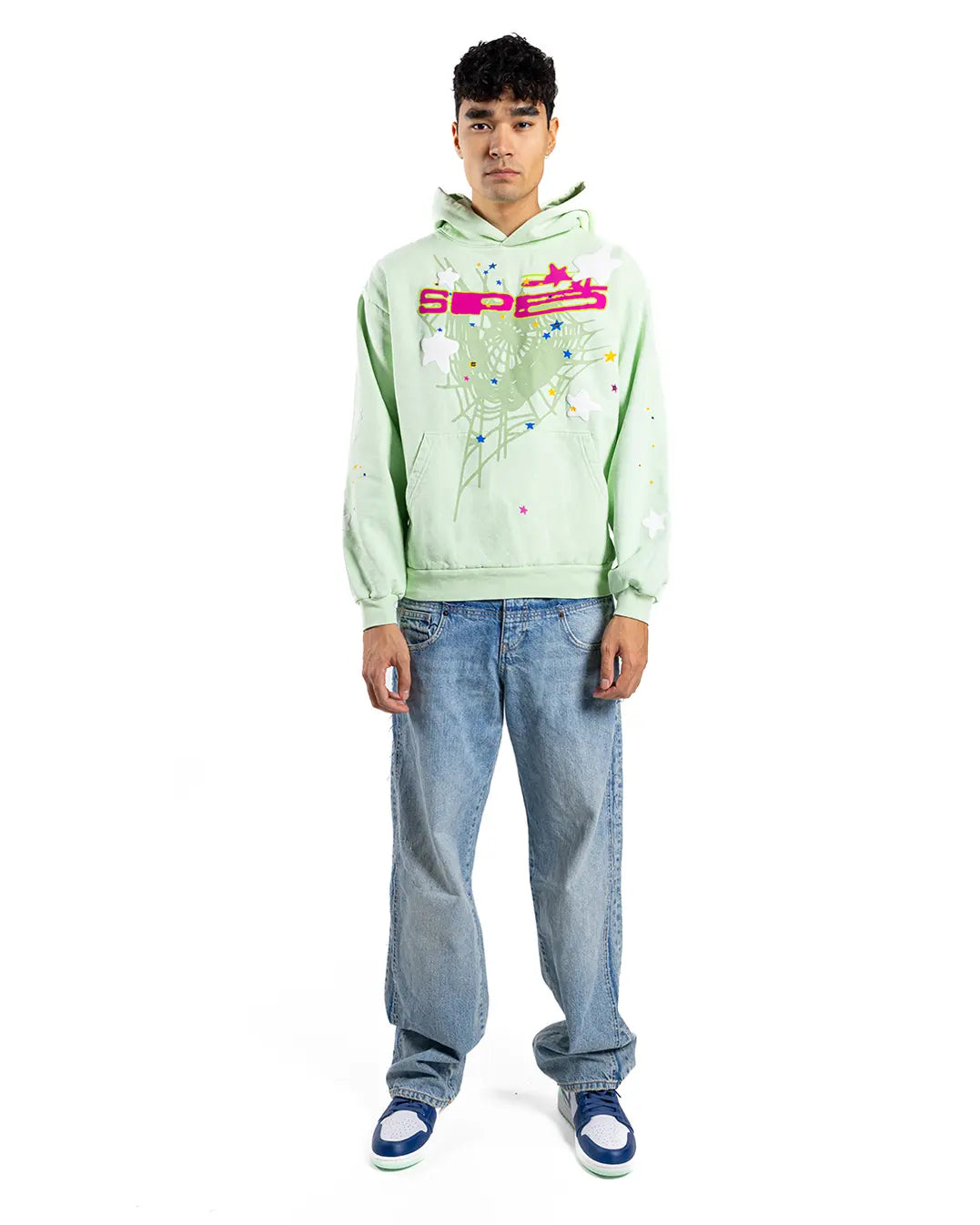 A young man stands facing forward in a Sp5der "Mint Star Web" Hoodie—mint green, 440GSM heavyweight oversized pullover—styled with loose blue jeans and blue-and-white sneakers, exemplifying Y2K streetwear against a white background.