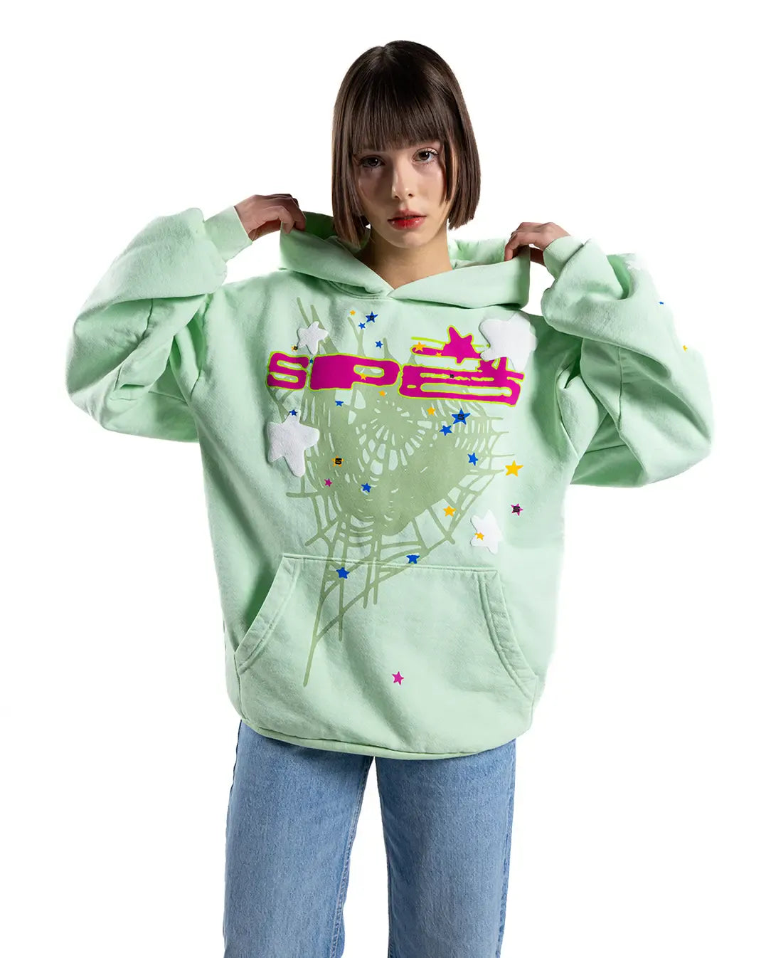 A woman wears a Sp5der “Mint Star Web” Hoodie—mint green, 440GSM heavyweight and oversized streetwear pullover—with colorful star and web designs, paired with blue jeans, posing against a plain white background.