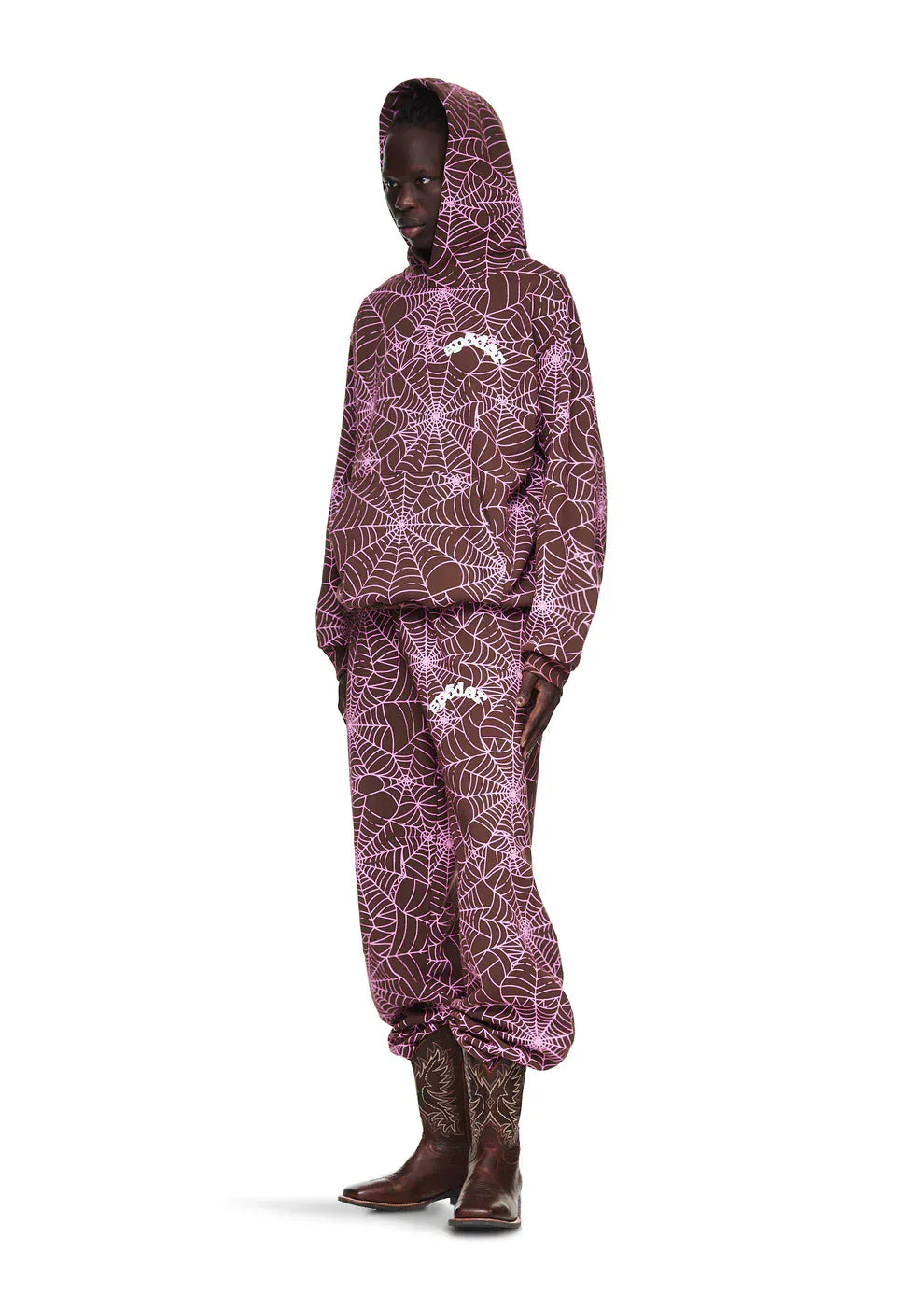 Wearing a Brown All-Over Spider Web Graphic Hoodie – Comfort Fit Pullover Hoodie with matching pants and brown cowboy boots, the person stands against a white background.