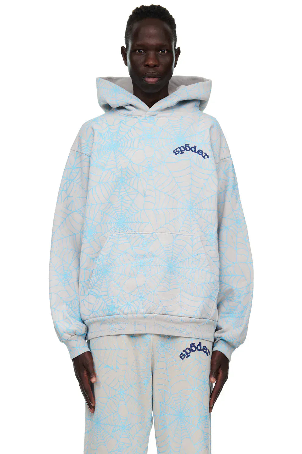 A person wears the Light Grey All-Over Spider Web Graphic Hoodie – Comfort Fit Pullover Hoodie, featuring blue spider web prints and Sp5der text for a bold, graphic look, paired with matching pants.