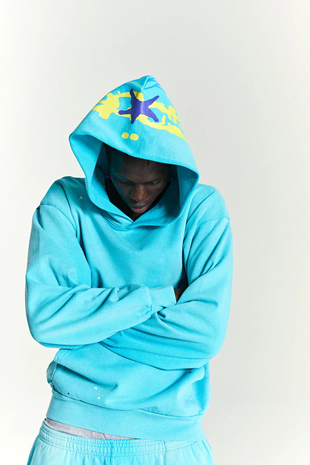Person wearing the Turquoise Graphic Hoodie with Star Detail and Spider Web Back Print, arms crossed and head lowered against a plain background.