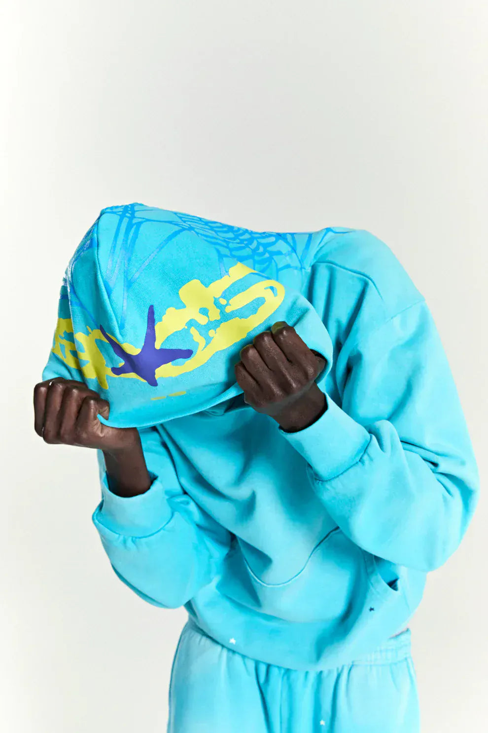 A person pulls the Turquoise Graphic Hoodie with Star Detail and Spider Web Back Print – Comfort Fit Pullover Hoodie over their head, obscuring their face, against a plain light background.