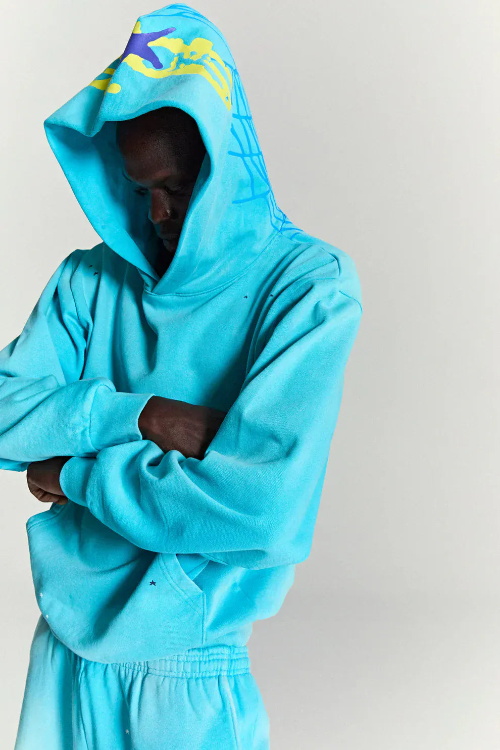 Person wearing a Turquoise Graphic Hoodie with Star Detail and Spider Web Back Print – a comfort fit pullover with unique graphic elements, standing with arms crossed and looking down.