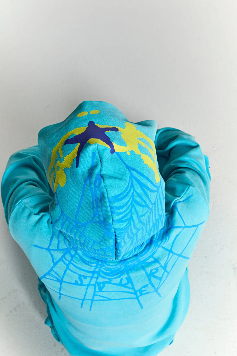 A person viewed from above wears the Turquoise Graphic Hoodie with Star Detail and Spider Web Back Print – a comfort fit pullover set against a light background.