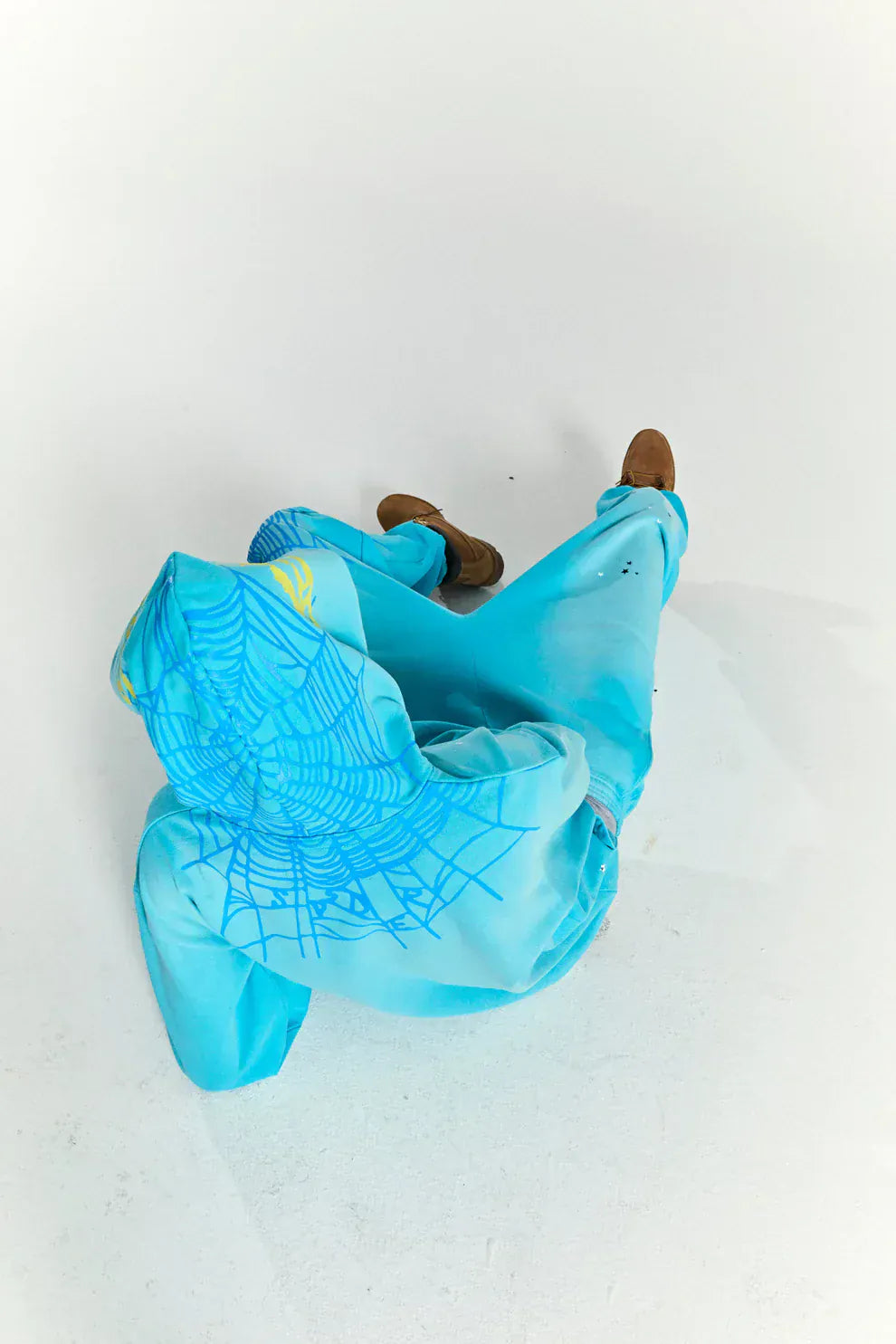 Person wearing a Turquoise Graphic Hoodie with Star Detail and Spider Web Back Print – Comfort Fit Pullover Hoodie and matching blue pants, sitting on a white floor with brown shoes.