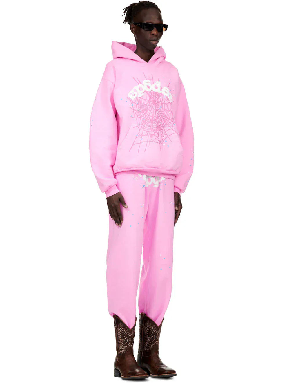 A person wears the Pink Graphic Hoodie with Spider Web Print – Comfort Fit Pullover Hoodie, paired with matching pink jagged hem pants, brown cowboy boots, and sunglasses, standing against a white background.