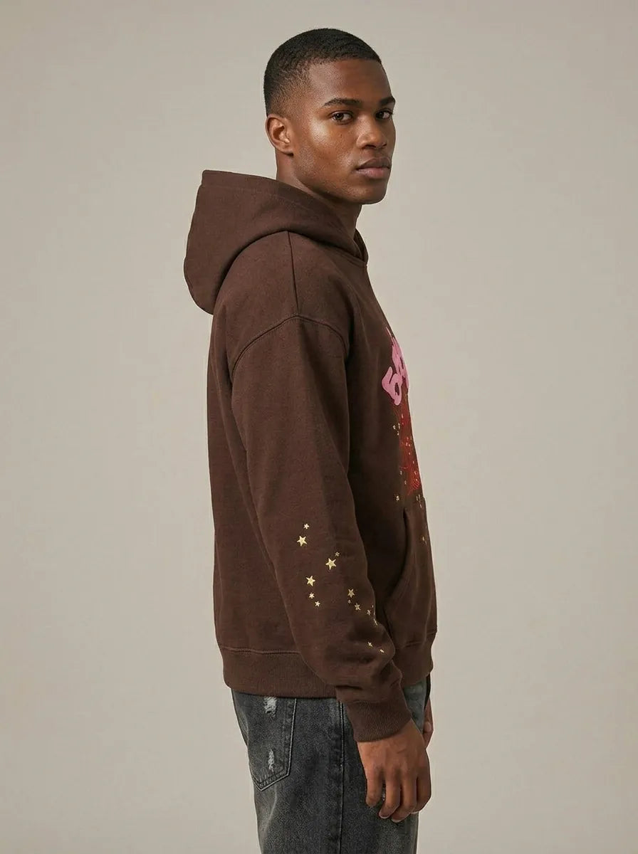 A man stands sideways in the 555 Brown Spider Hoodie – Sp5der 555 Web Brown/Pink, a heavyweight oversized pullover with a pink chest graphic and gold star sleeve accents, paired with dark jeans against a plain background.