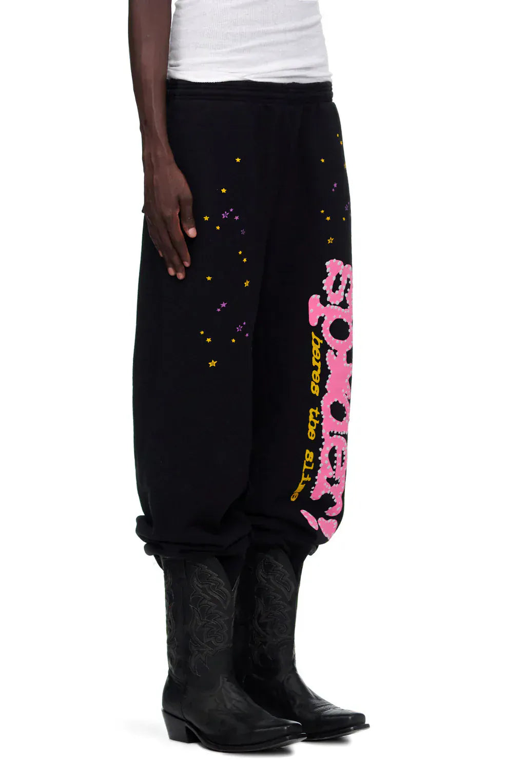 A person wearing Sp5der Sweatpants – unisex black cotton sweatpants with pink graphic logo and star print – pairs them with black cowboy boots and a white tank top for a bold streetwear look.